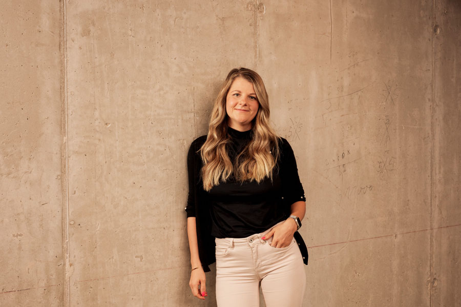 Eine Person lehnt an einer Betonwand, trägt ein schwarzes Oberteil und helle Hosen.