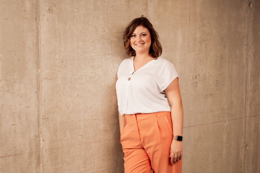 Eine Frau in weißem Oberteil und orangefarbener Hose lehnt an einer Betonwand und lächelt.