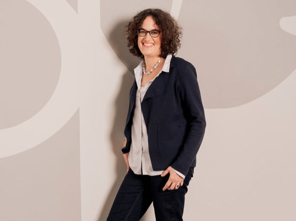 Eine Frau mit Brille und lockigem Haar trägt Blazer, Bluse und Jeans vor einer grauen Wand mit weißen Mustern.