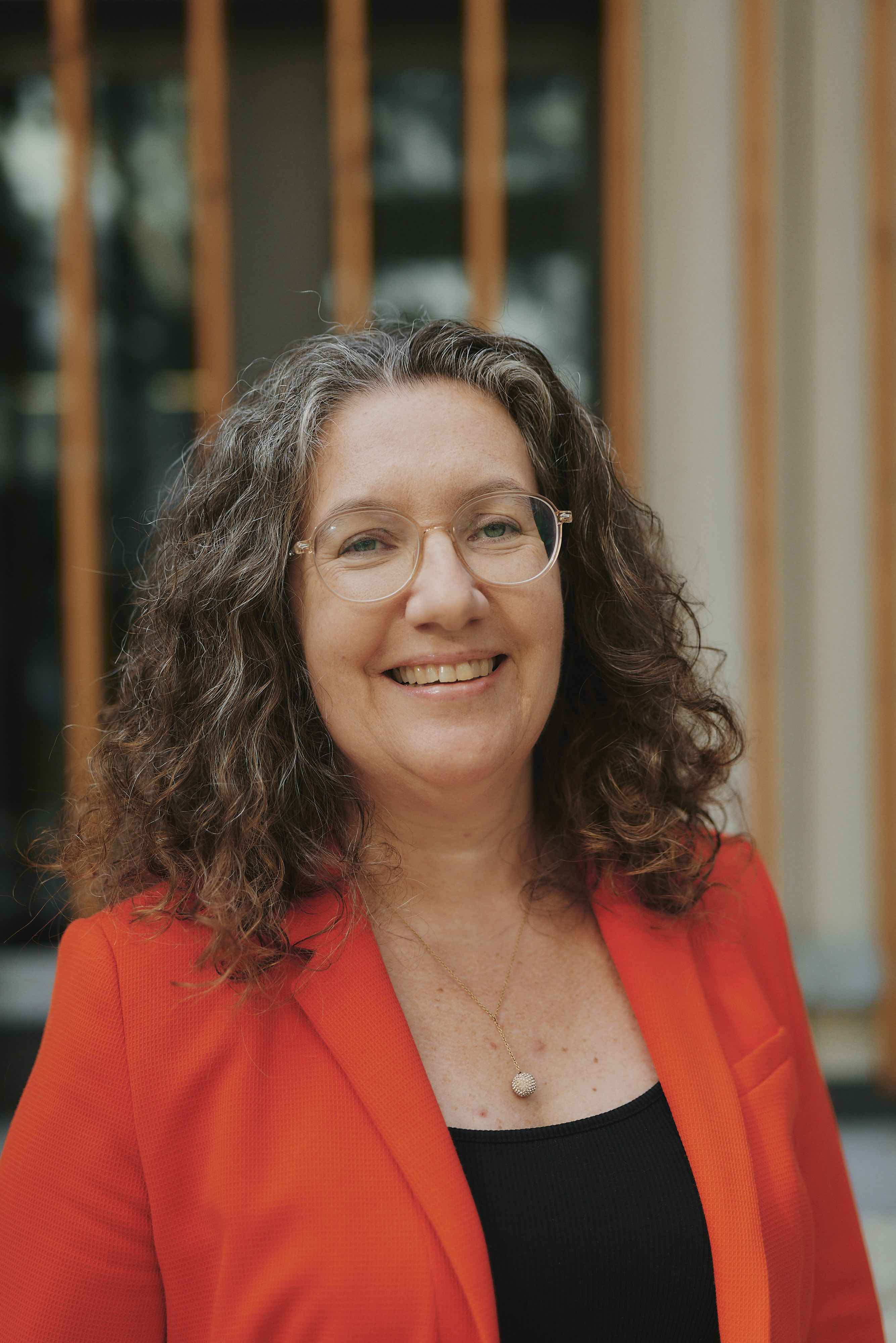 Eine lächelnde Person mit lockigem Haar trägt eine Brille und einen roten Blazer.