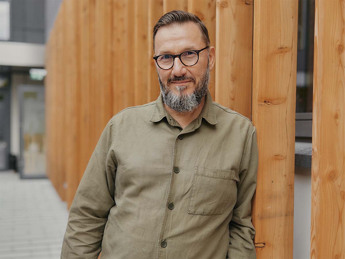 Ein Mann mit Brille lehnt an einer Holzlattenwand und trägt ein olivgrünes Hemd.