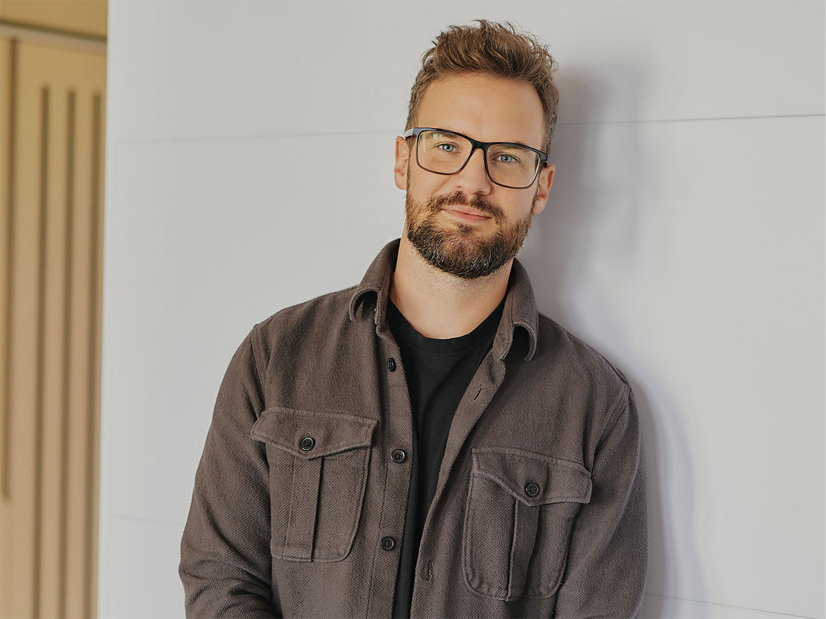 Ein Mann mit Brille und Bart lehnt lächelnd an einer weißen Wand, trägt ein braunes Hemd.