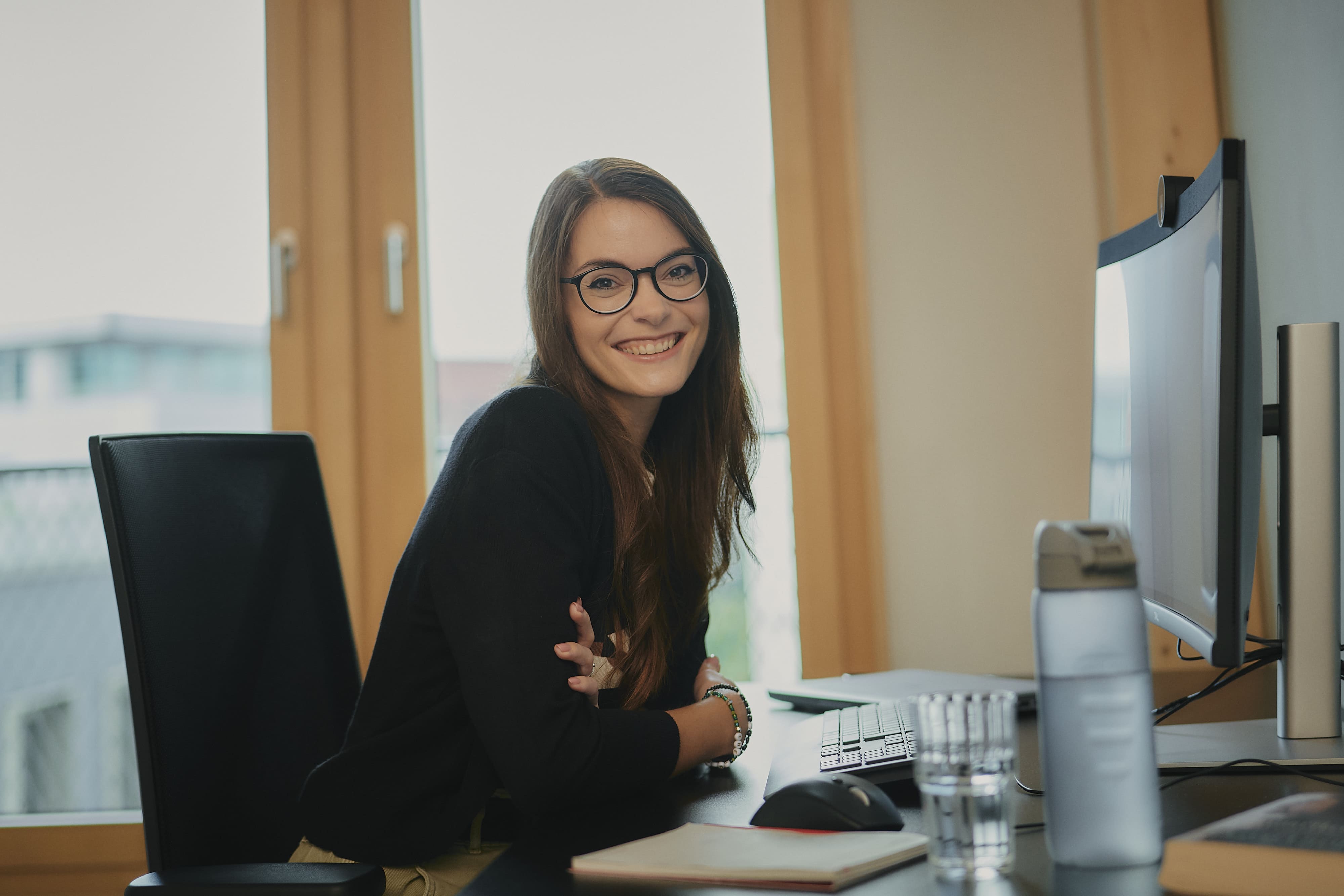 Frau mit Brille lächelt am Schreibtisch vor einem Monitor, neben ihr eine Wasserflasche und Notizbuch.