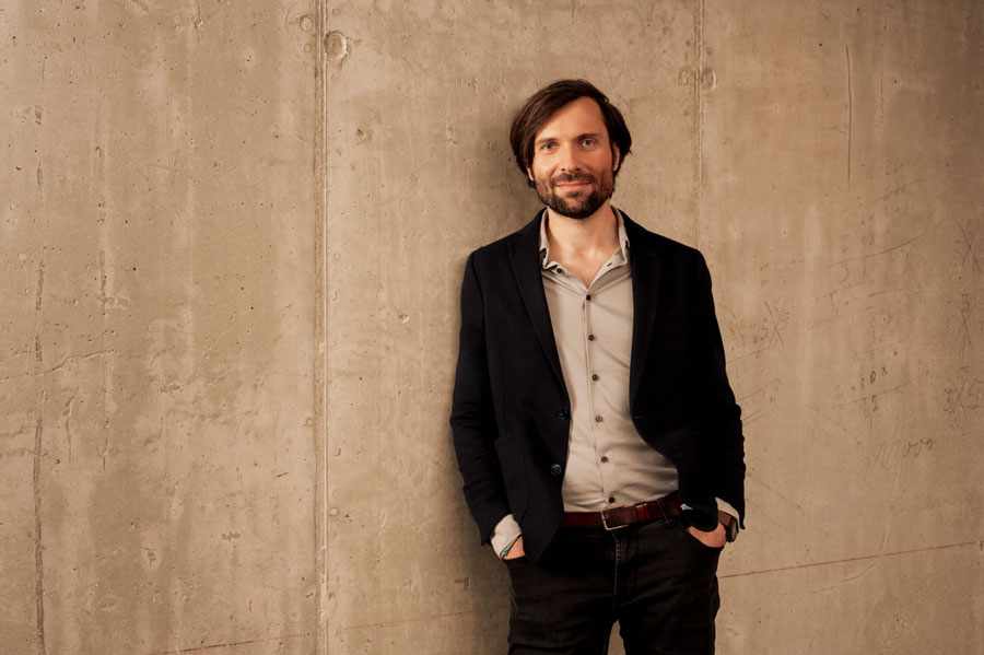 Ein Mann in schwarzem Blazer lehnt lächelnd an einer Betonwand.