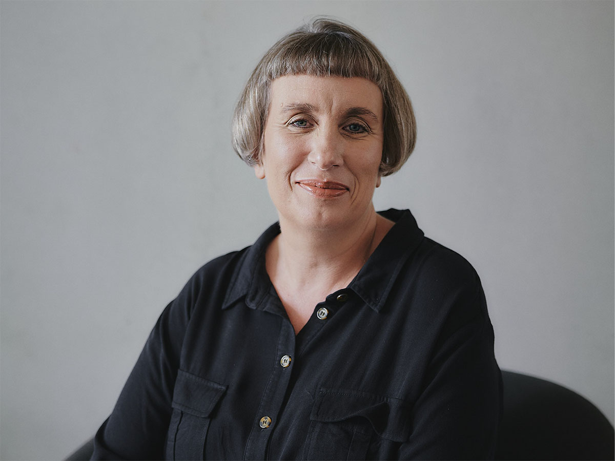 Eine Person mit kurzem Haar lächelt in die Kamera, trägt ein dunkles Hemd vor grauem Hintergrund.