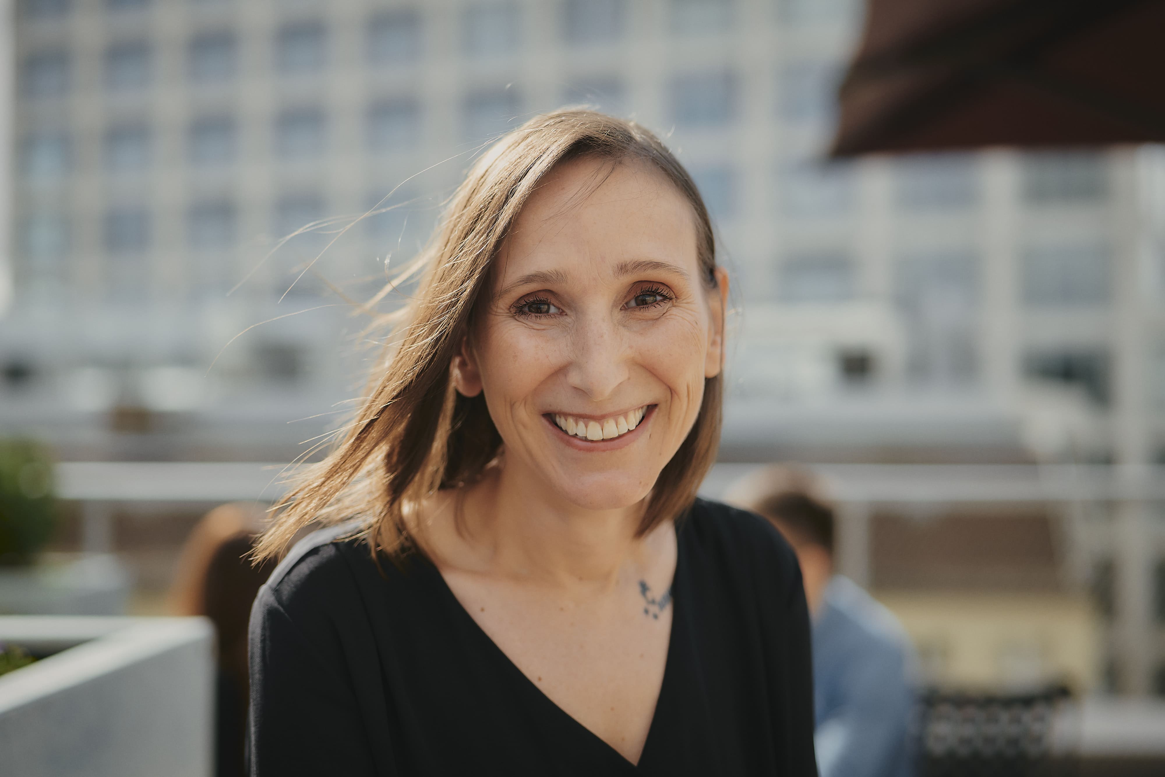Eine lächelnde Frau mit schulterlangem Haar steht draußen vor einem unscharfen Gebäude.