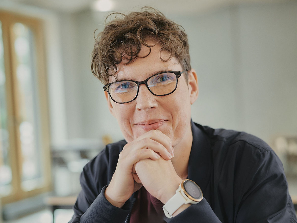 Eine Person mit Brille lächelt, stützt das Kinn auf gefaltete Hände, unscharfer Hintergrund.