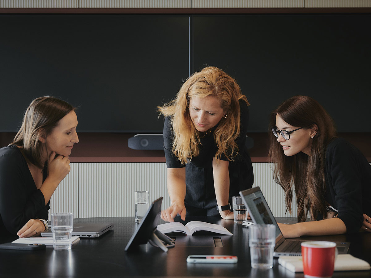 Drei Frauen diskutieren an einem Konferenztisch mit Laptops und Dokumenten.