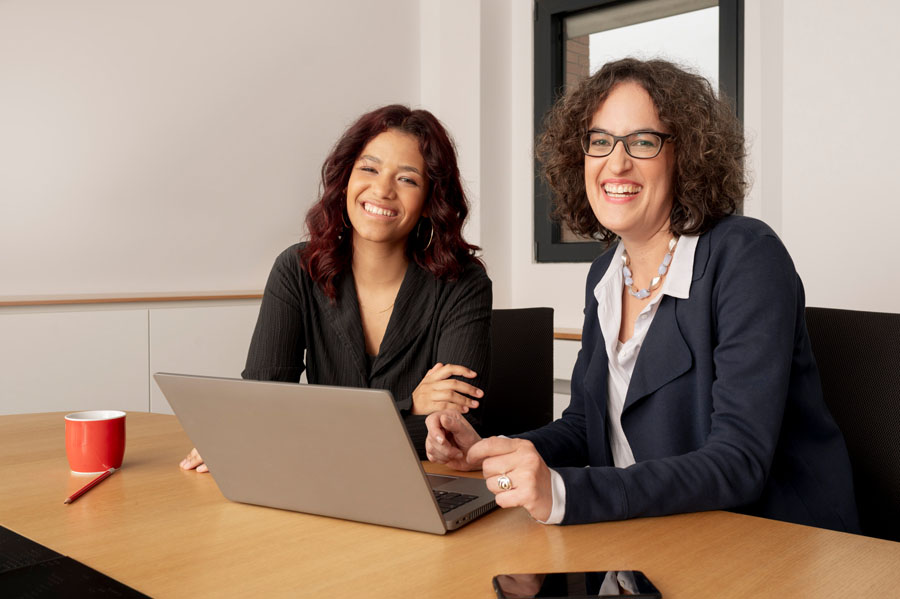 Zwei lächelnde Frauen sitzen am Tisch mit Laptop und roter Tasse.