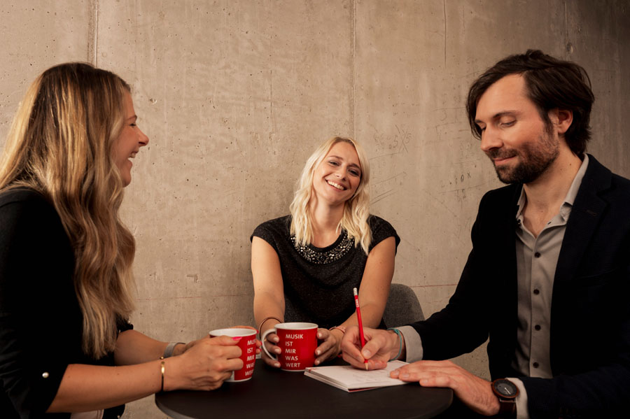 Drei Personen lachen und sitzen an einem Tisch mit roten Tassen vor einer Betonwand.