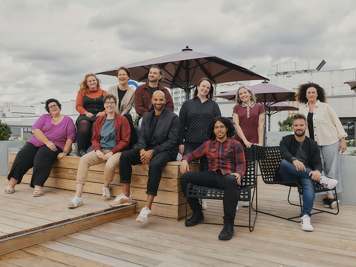 Eine Gruppe von zwölf Personen sitzt und steht auf einer Terrasse mit Holzboden und Sonnenschirmen.