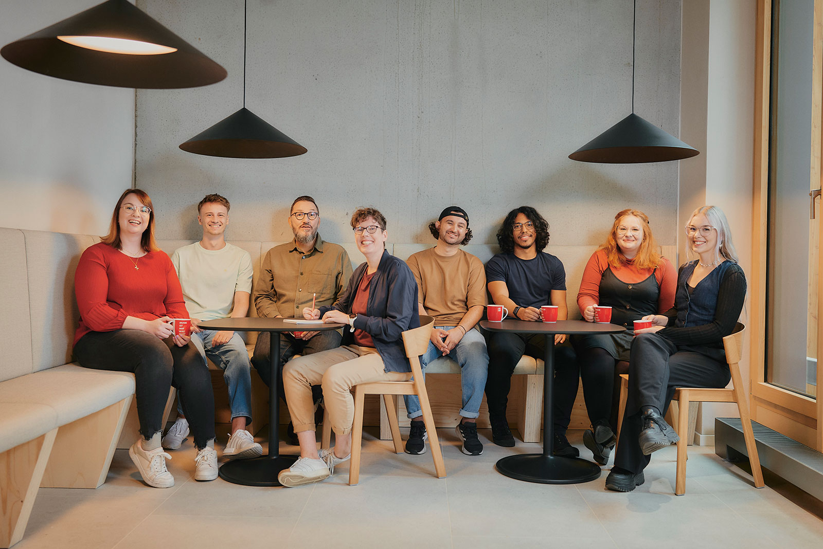Eine Gruppe von acht Personen sitzt lächelnd an einem Tisch mit roten Tassen in einem modernen Raum.