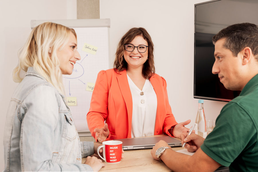Drei Personen diskutieren lächelnd an einem Tisch, Flipchart im Hintergrund. Eine Frau trägt einen roten Blazer.