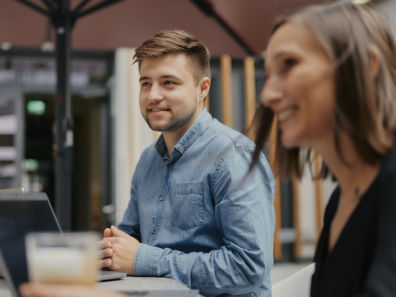 Zwei Personen lächeln und sitzen an einem Tisch mit einem Laptop im Freien.