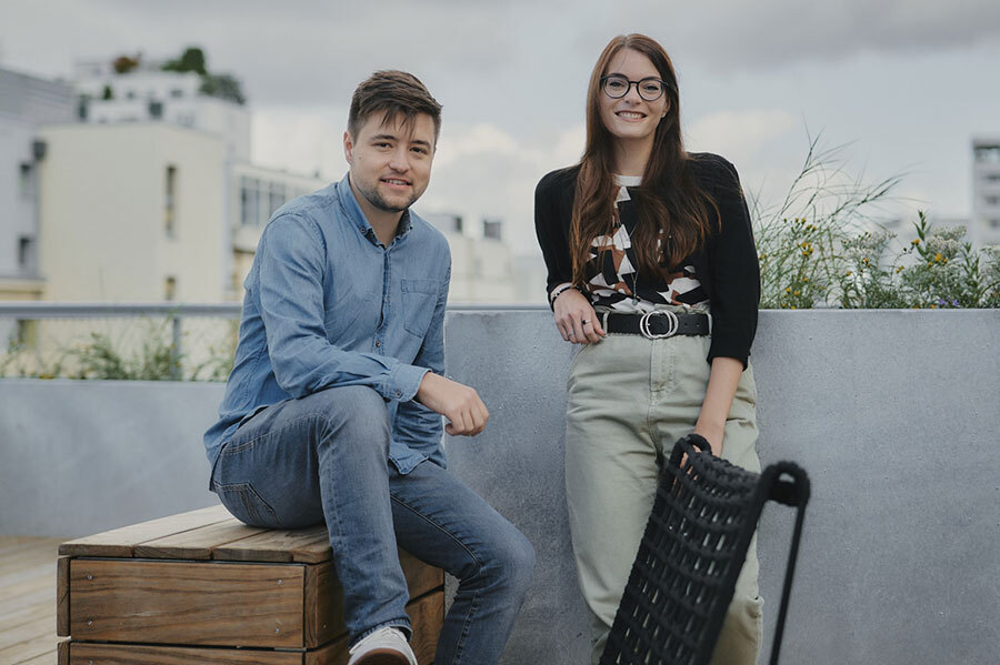 Zwei Personen auf einer Dachterrasse, eine sitzt auf einer Holzbank, die andere steht daneben, beide lächeln.