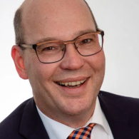 Ein lächelnder Mann mit Brille in Anzug und Krawatte.