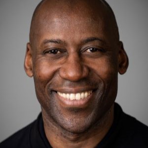 Ein lächelnder Mann in einem schwarzen Hemd vor einem grauen Hintergrund.