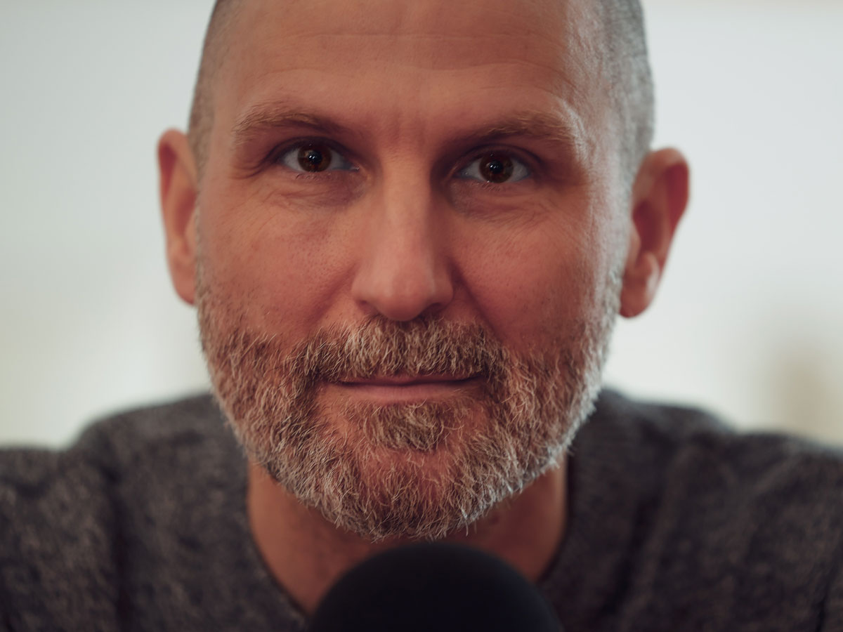 Porträt eines lächelnden Mannes mit Bart, grauem Pullover und Mikrofon im Vordergrund.