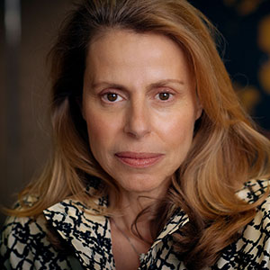 A woman with long, light brown hair wearing a patterned blouse, looking directly at the camera.