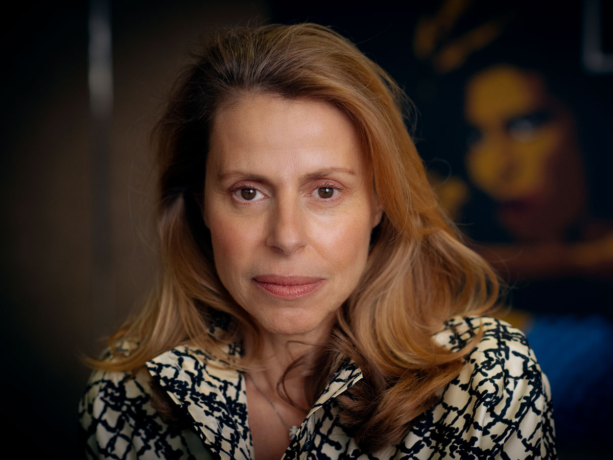 Eine Frau mit langen blonden Haaren schaut ernst in die Kamera, trägt ein gemustertes Oberteil.