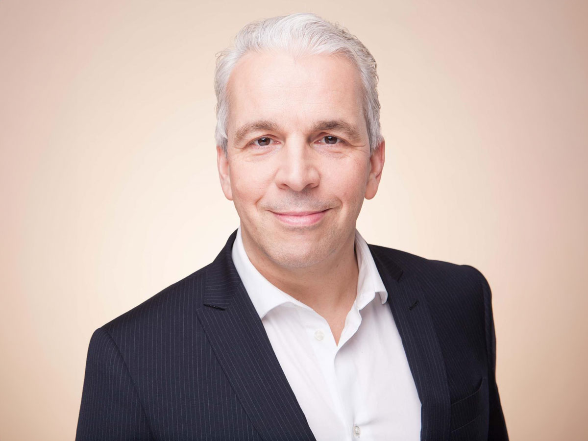 Ein Mann in Anzug und Hemd lächelt vor neutralem Hintergrund.