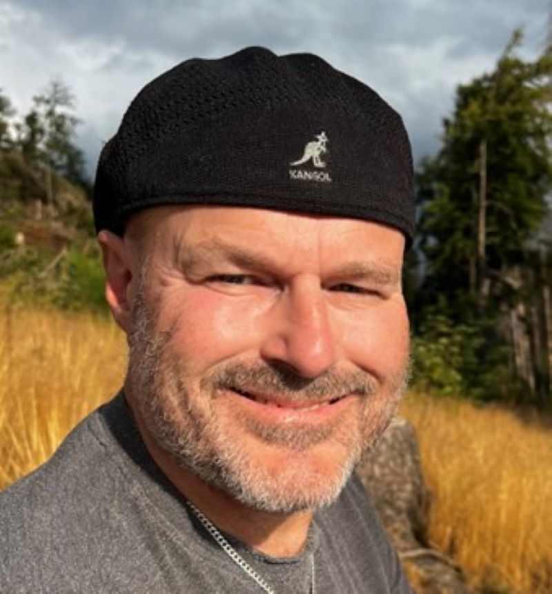 Ein Mann mit schwarzer Mütze lächelt im Freien, im Hintergrund sind Bäume und gelbes Gras.