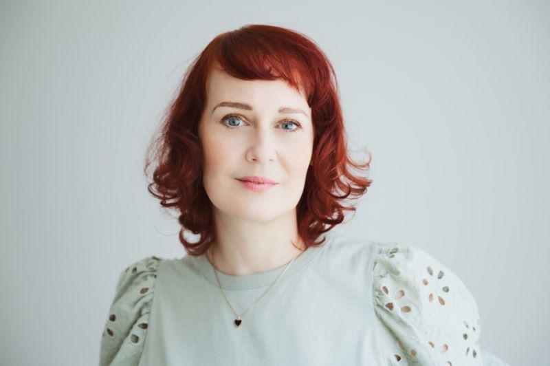 Eine Frau mit roten Haaren trägt ein helles Oberteil vor grauem Hintergrund.