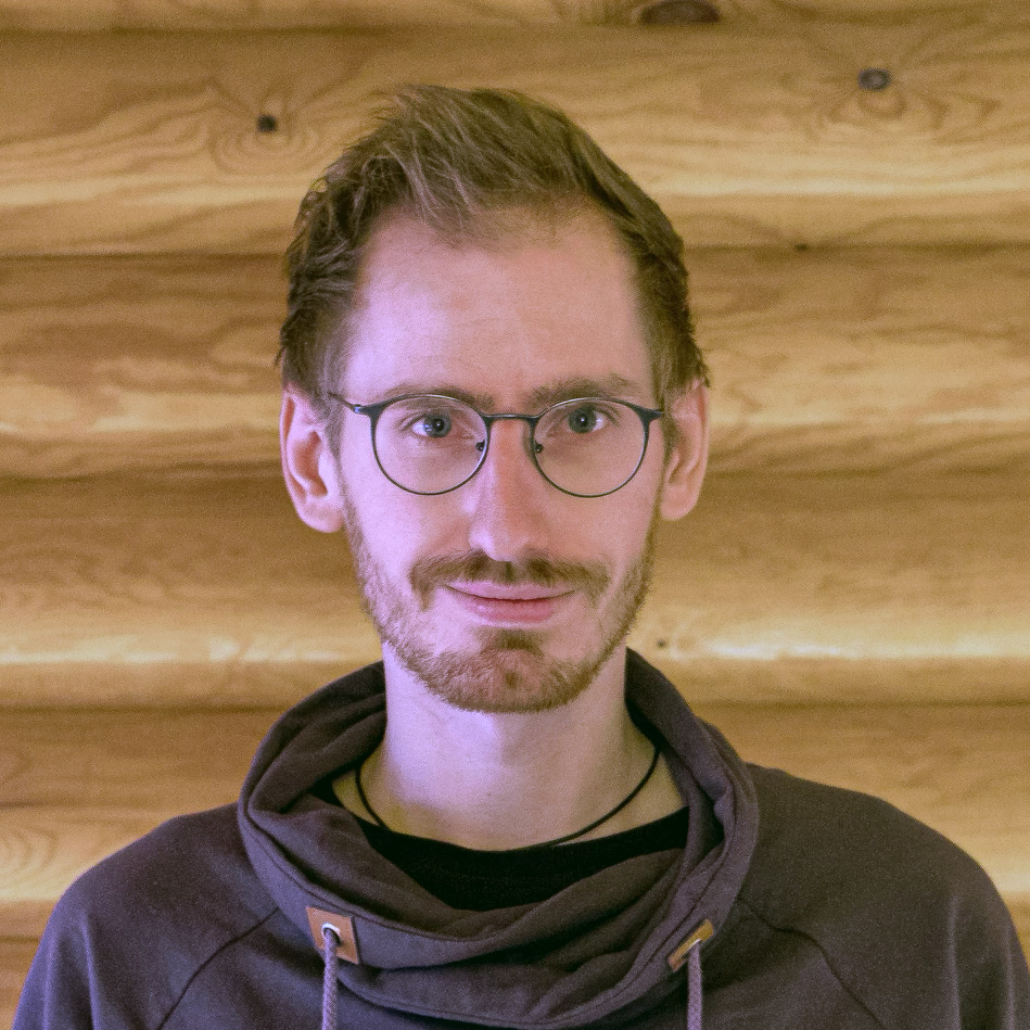 Ein Mann mit Brille vor einer Holzwand, trägt einen dunkelgrauen Kapuzenpullover.