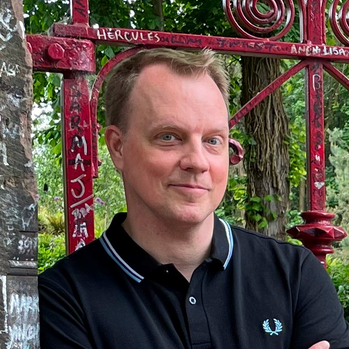 Eine Person steht vor einem roten, verzierten Tor in einem grünen Garten.