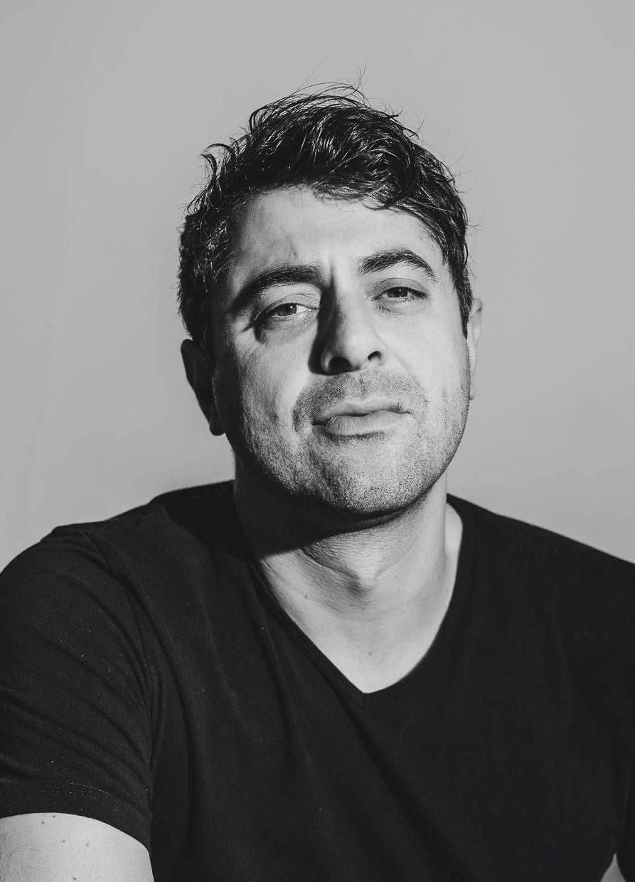 Schwarz-weiß Porträt eines Mannes mit dunklem Haar und Bart, in einem schwarzen T-Shirt, schaut ernst in die Kamera.