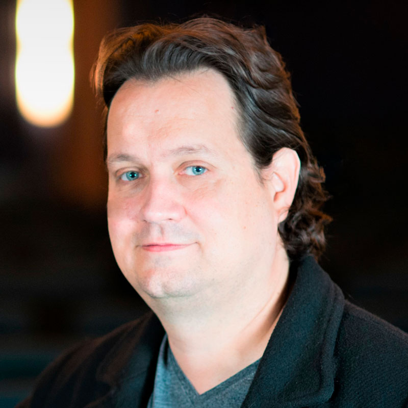 Ein Mann mit braunen Haaren und blauer Jacke lächelt leicht in die Kamera vor dunklem Hintergrund.