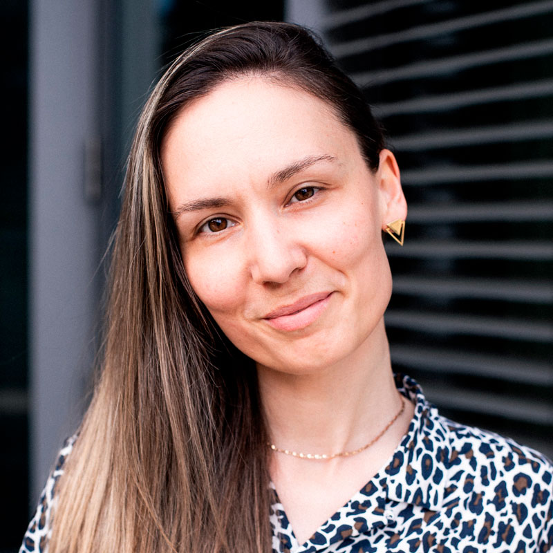 Eine Frau lächelt in die Kamera, trägt ein gemustertes Oberteil und goldene Ohrringe.