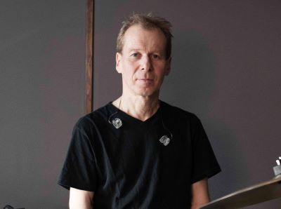 Eine Person in schwarzem T-Shirt steht vor einer grauen Wand und trägt In-Ear-Kopfhörer.