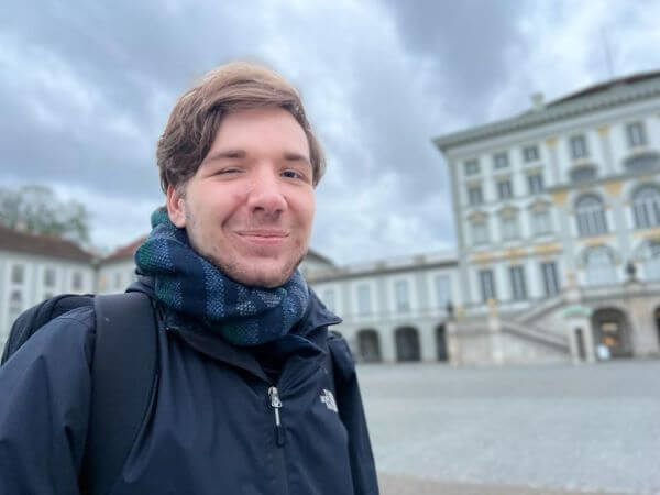 Eine Person in dunkler Jacke steht vor einem historischen Gebäude unter bewölktem Himmel.