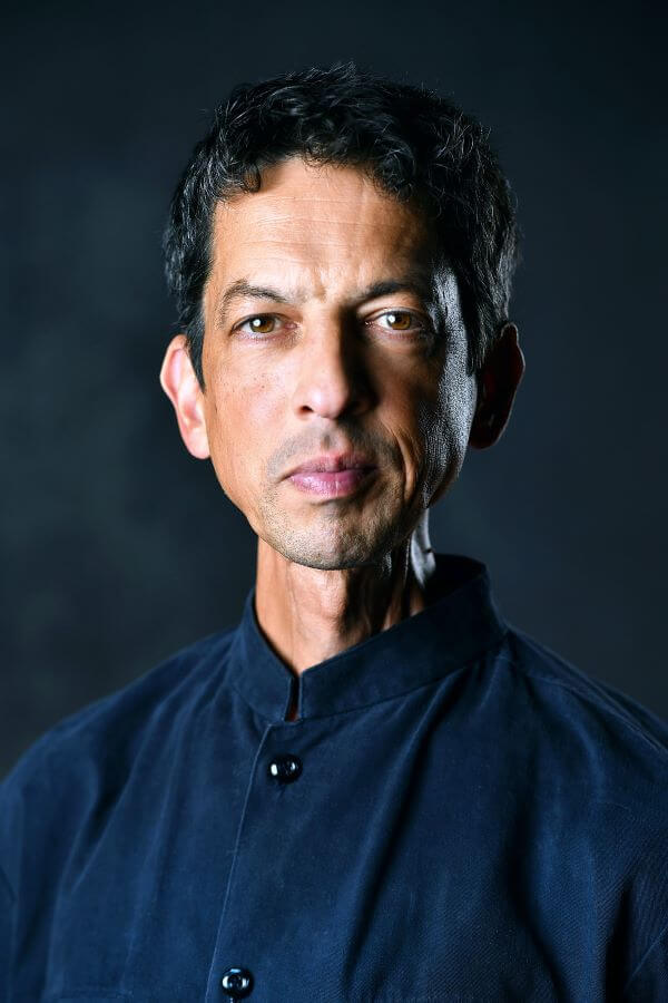 Ein Mann in dunklem Hemd vor dunklem Hintergrund, schaut ernst in die Kamera.