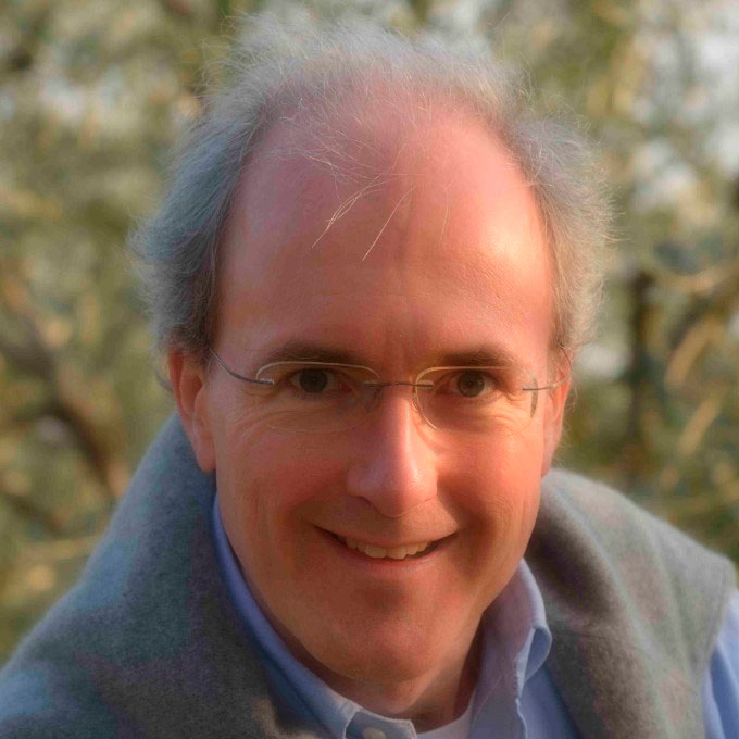 Ein lächelnder Mann mit Brille und grauem Haar vor unscharfem, grünem Hintergrund.