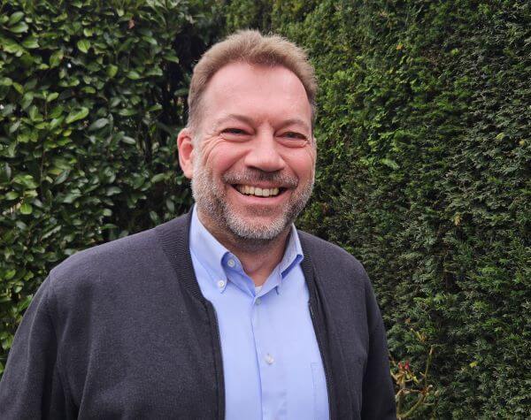 Ein lächelnder Mann in Hemd und Pullover steht vor einer grünen Hecke.