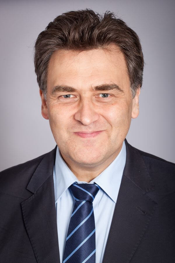 Ein Mann in Anzug und Krawatte lächelt vor neutralem Hintergrund.