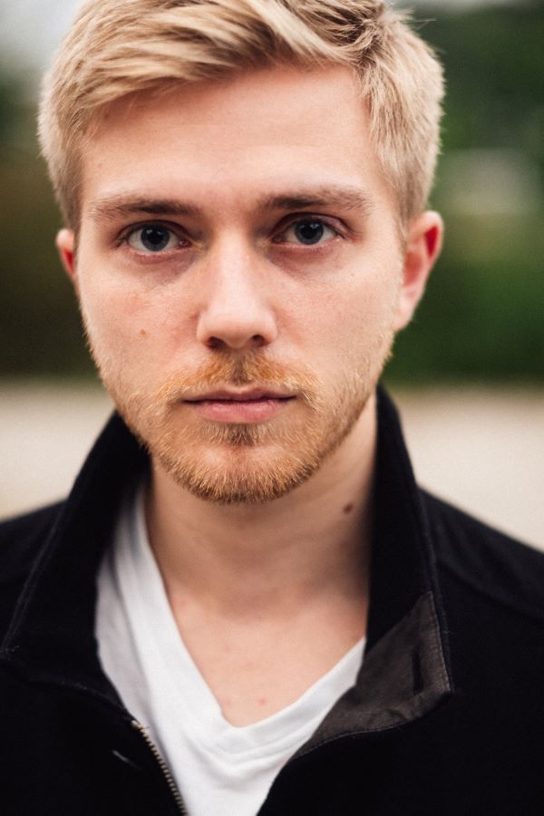 Ein junger Mann mit blondem Haar und Bart schaut direkt in die Kamera, trägt eine schwarze Jacke und ein weißes T-Shirt.
