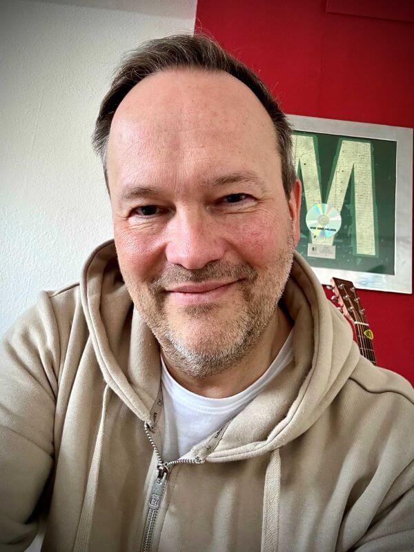 Ein Mann in beigem Hoodie lächelt vor einer roten Wand mit gerahmtem Bild und Gitarre im Hintergrund.