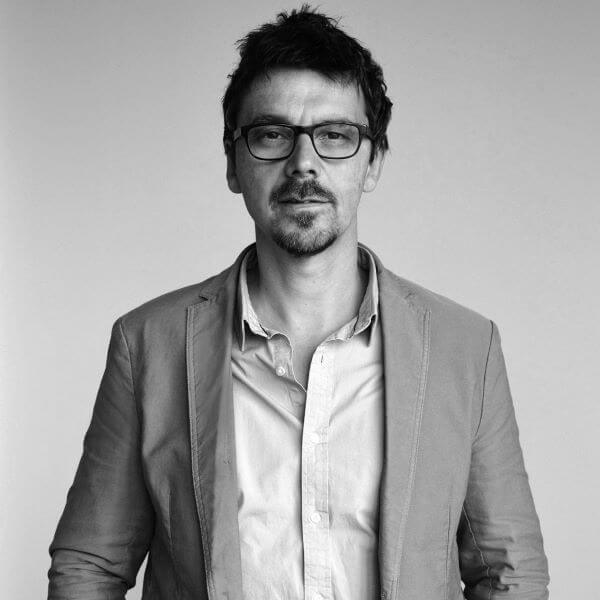 Ein Mann mit Brille und Bart in einem Sakko vor neutralem Hintergrund.