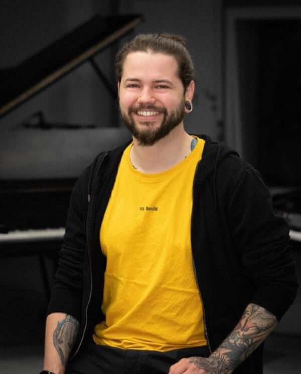 Ein lächelnder Mann mit Bart in gelbem Shirt und schwarzer Jacke sitzt vor einem Klavier.