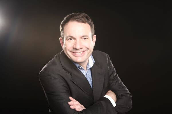 Ein lächelnder Mann in Anzug posiert mit verschränkten Armen vor schwarzem Hintergrund.