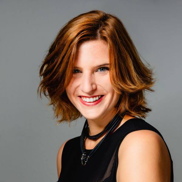 Eine lächelnde Frau mit kinnlangem, rotem Haar vor grauem Hintergrund.