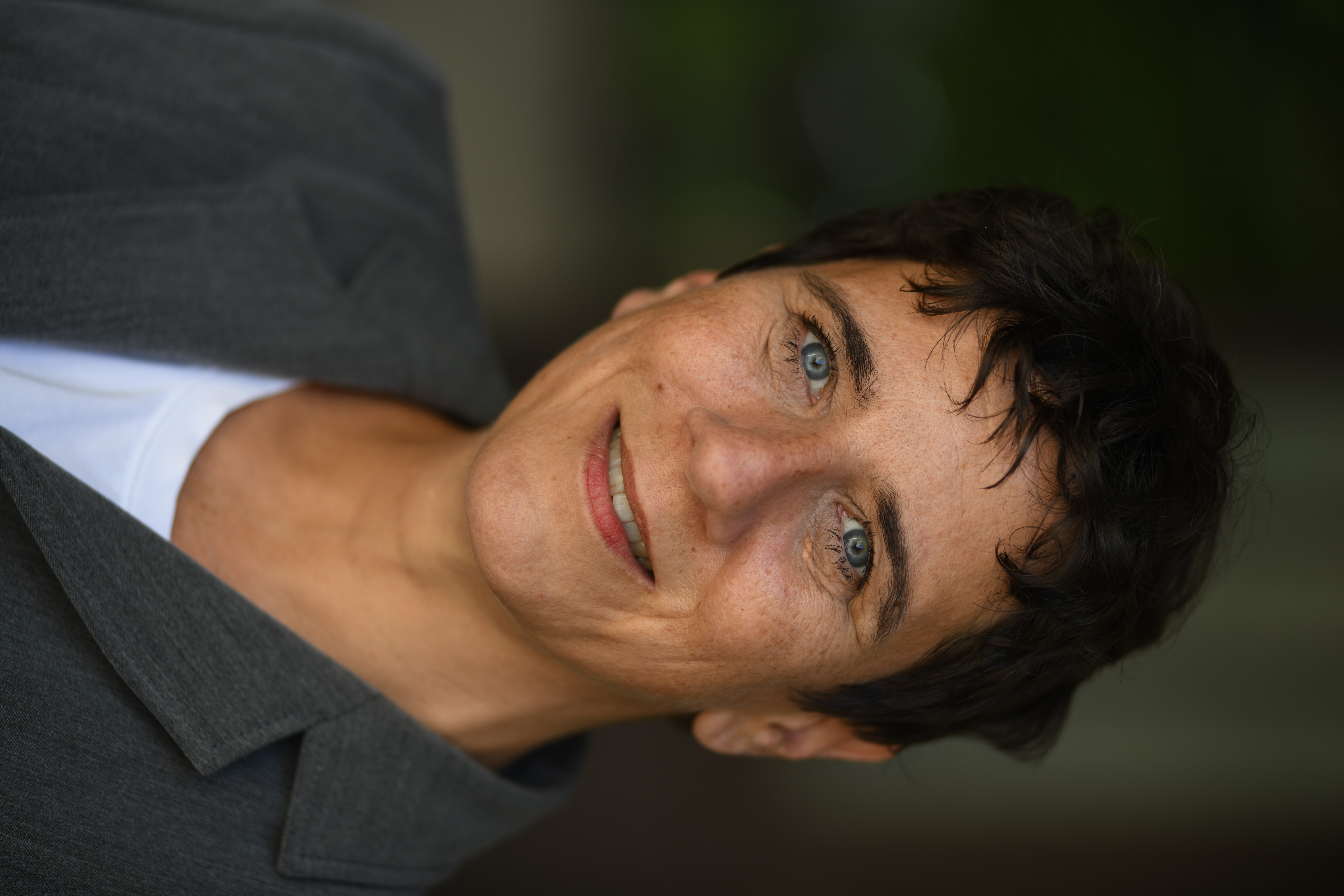 Eine lächelnde Person in grauem Blazer mit kurzen dunklen Haaren vor unscharfem Hintergrund.