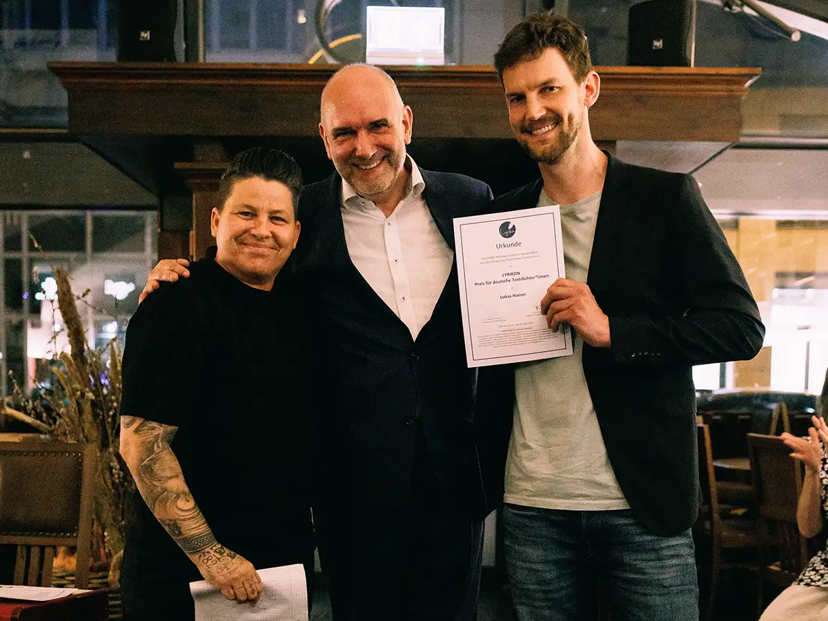 Drei Personen lächeln in die Kamera, eine Person hält eine Urkunde in einem Innenraum.