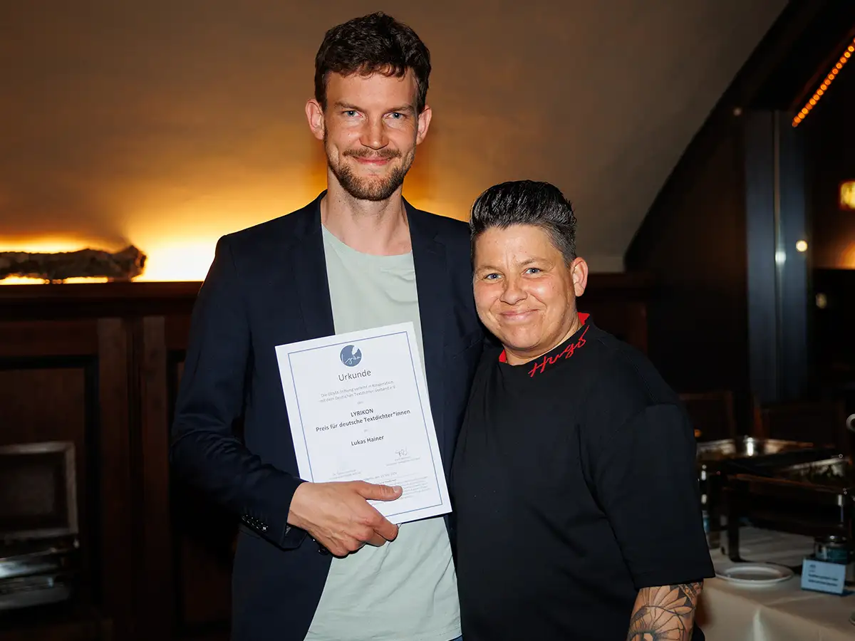 Zwei Personen lächeln in die Kamera, eine hält eine Urkunde in der Hand. Hintergrund mit warmer Beleuchtung.