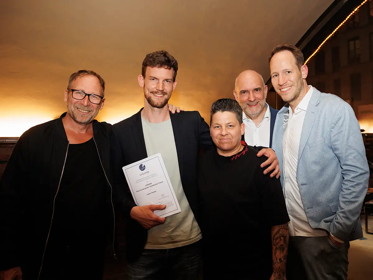 Fünf lächelnde Personen posieren zusammen, eine hält eine Urkunde in der Hand, in einem beleuchteten Raum.