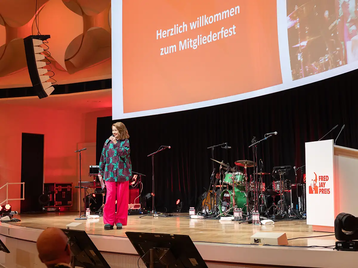 Eine Frau spricht auf einer Bühne mit Instrumenten, roter Hintergrund, Text: 