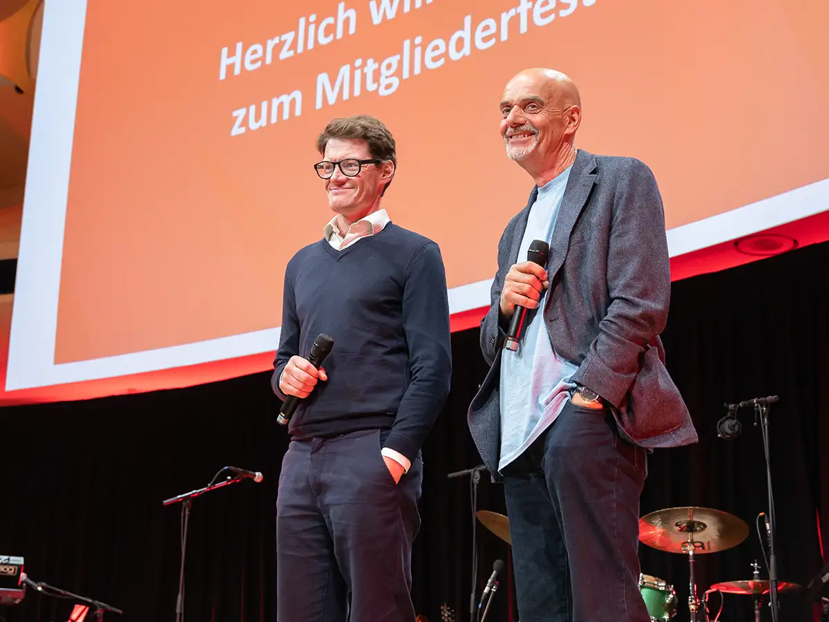 Zwei Männer mit Mikrofonen auf einer Bühne vor einem roten Hintergrund mit Begrüßungstext.